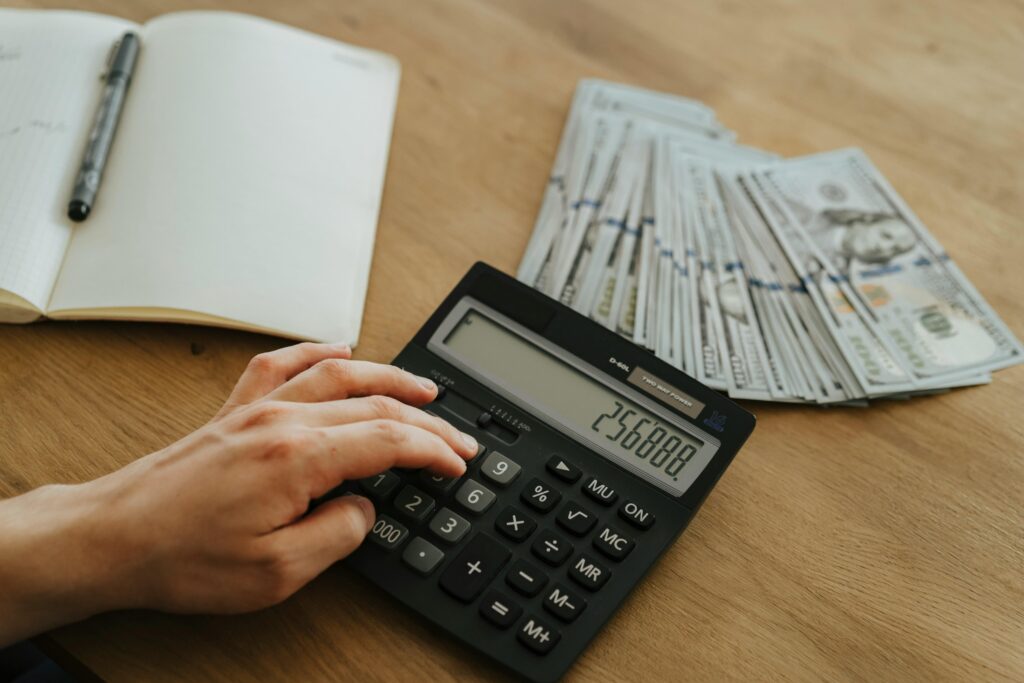pexels photo 5466806 5466806 A hand calculates financial figures using a calculator with stacks of cash nearby on a wooden table.